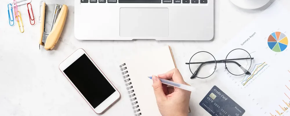 Ein Schreibtisch mit Laptop, Smartphone, Brille, Kreditkarte, einer Pflanze, einem Tacker und einem Schreibblock auf dem eine Hand zum Schreiben ansetzt